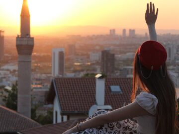 A woman in traditional attire waves at the city skyline during a stunning sunset.