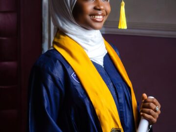 Portrait of a smiling young Muslim woman in a graduation cap and gown holding a diploma.