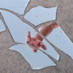 A hand reaching out is reflected in scattered pieces of a broken mirror on a textured surface.