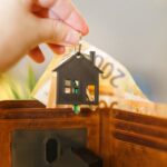 A hand holds a house-shaped keychain above an open wallet with Euro banknotes.