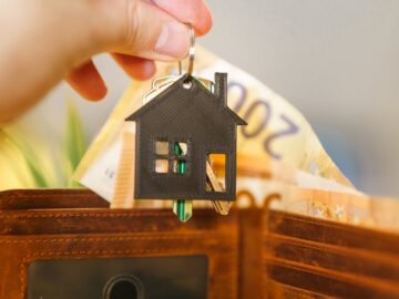 A hand holds a house-shaped keychain above an open wallet with Euro banknotes.