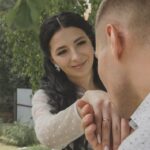 A woman smiles as a man kisses her hand outdoors, symbolizing love and commitment.