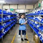Female worker organizing inventory in a modern warehouse aisle, surrounded by blue storage bins.
