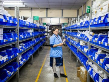 Female worker organizing inventory in a modern warehouse aisle, surrounded by blue storage bins.