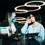 Two women in a modern office discussing with digital devices, highlighting contemporary teamwork.