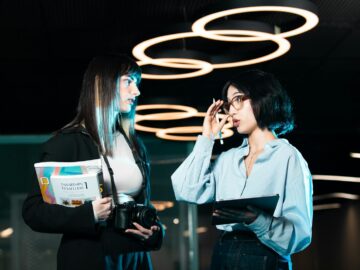 Two women in a modern office discussing with digital devices, highlighting contemporary teamwork.