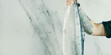 A tattooed man holds a large fish by the tail against a marble background. Perfect for culinary or seafood themes.