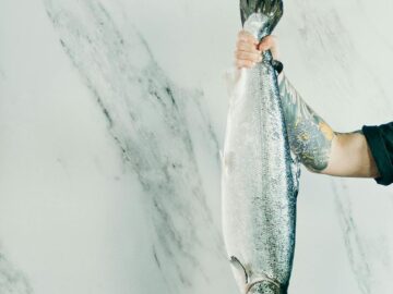 A tattooed man holds a large fish by the tail against a marble background. Perfect for culinary or seafood themes.