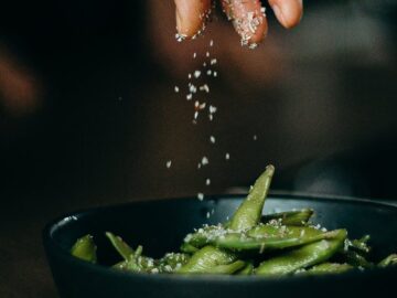 A hand sprinkles salt on fresh edamame, showcasing a delicious and healthy cooking moment.