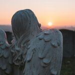 A contemplative angel statue in a cemetery, facing a serene sunset sky.