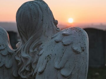 A contemplative angel statue in a cemetery, facing a serene sunset sky.