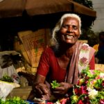 Elderly flower vendor smiling warmly at outdoor market stall, selling fresh blossoms.