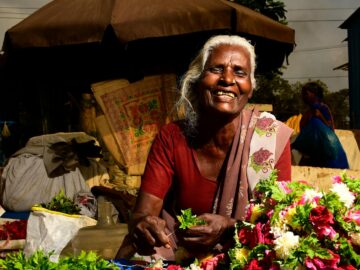 Elderly flower vendor smiling warmly at outdoor market stall, selling fresh blossoms.