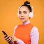 A young woman listens to music on wireless headphones while using a smartphone against an orange background.
