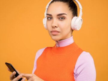 A young woman listens to music on wireless headphones while using a smartphone against an orange background.