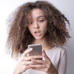 A young woman with curly hair looks surprised while reading something on her smartphone indoors.