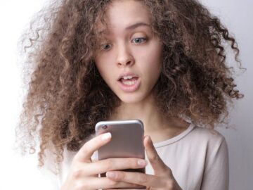 A young woman with curly hair looks surprised while reading something on her smartphone indoors.