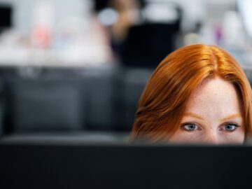 A woman with red hair intensely focused on a computer screen in an office setting.