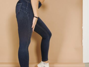 Side view of crop faceless sporty blond lady in tight dark blue jeans and white sneakers standing leaned on one leg near beige wall