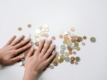 Hands arranging a variety of international coins on a neutral light background.