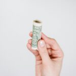 A hand holding a rolled US dollar bill against a plain background, symbolizing finance and savings.