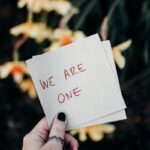 A hand holding a note reading 'We Are One' against a floral outdoor backdrop.