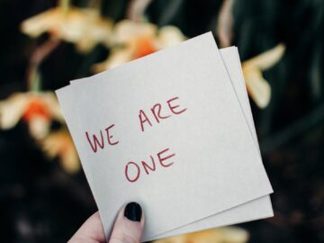 A hand holding a note reading 'We Are One' against a floral outdoor backdrop.