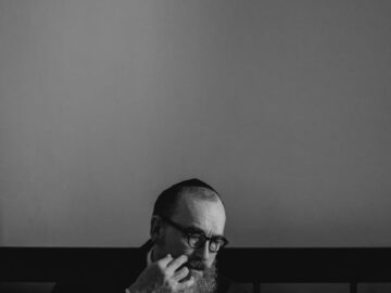 An Orthodox Jewish man with a beard and glasses, reading a book indoors.