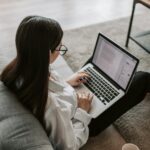 Woman working remotely with a laptop on the floor next to a sofa, enjoying comfortable home office setup.