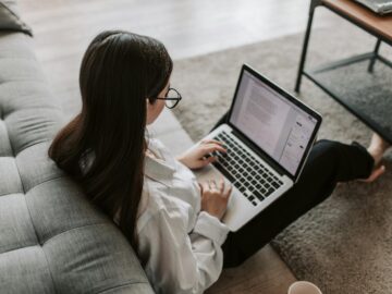 Woman working remotely with a laptop on the floor next to a sofa, enjoying comfortable home office setup.