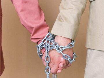 Close-up of two individuals holding hands bound by chains, symbolizing connections and relationships.