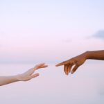 Two diverse hands reaching towards each other over a serene pastel sky and water background.