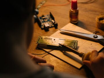 Person soldering a printed circuit board at a workstation with tools and components.