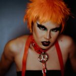 Striking portrait of a model with vibrant orange hair and dramatic makeup in a creative studio setting.