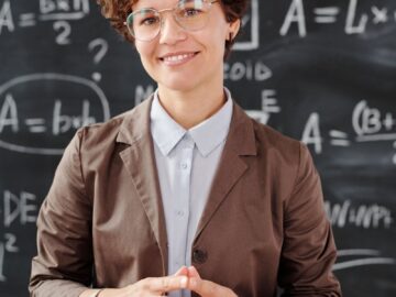 Teacher standing by a blackboard with algebra equations, smiling confidently.