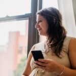 Smiling pregnant woman using smartphone near window, enjoying a peaceful moment indoors.
