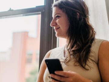 Smiling pregnant woman using smartphone near window, enjoying a peaceful moment indoors.