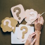 Hands holding pen next to paper question marks on notebook, symbolizing inquiry and creativity.