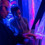 Two individuals working on laptops in a neon-lit setting, emphasizing technology and teamwork.