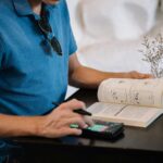 Adult man using smartphone and reading a book at a table indoors.