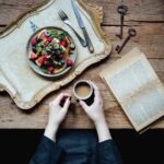 Top view of anonymous female standing near wooden table with book and cup of coffee with homemade pancakes topped with fruits and berries served on tray