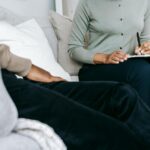 Crop anonymous female psychologist writing in clipboard and talking to black male client during psychotherapy appointment in light studio