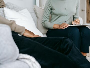 Crop anonymous female psychologist writing in clipboard and talking to black male client during psychotherapy appointment in light studio