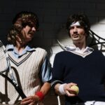 Two men in retro tennis attire with rackets and a tennis ball against a brick wall.