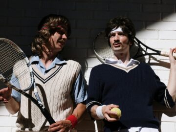 Two men in retro tennis attire with rackets and a tennis ball against a brick wall.