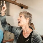 Senior caucasian woman attentively pointing during indoor seminar with diverse audience.