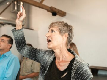 Senior caucasian woman attentively pointing during indoor seminar with diverse audience.