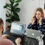 Two coworkers using laptops and phones while working together in a modern office setting.
