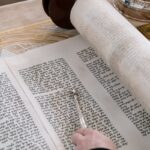 A detailed close-up of a Torah scroll with a yad pointer being used during a religious ceremony.