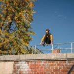 A young man executing a dynamic parkour jump over a brick wall in an urban setting, under a clear blue sky.